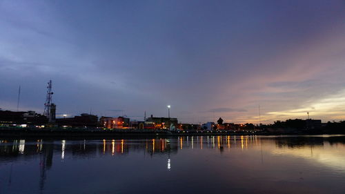 Illuminated buildings by river against sky at sunset