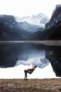 Distant unrecognizable female hiker with blanket in outstretched arms standing on shore of calm lake with smooth water surface reflecting rocky mountains covered with snow in overcast autumn day