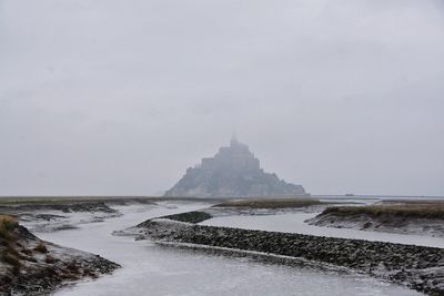 Stream flowing by mont saint-michel against sky during foggy weather