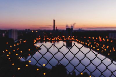 Fence by illuminated city against sky during sunset