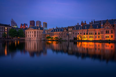 Illuminated buildings by river against blue sky at dusk