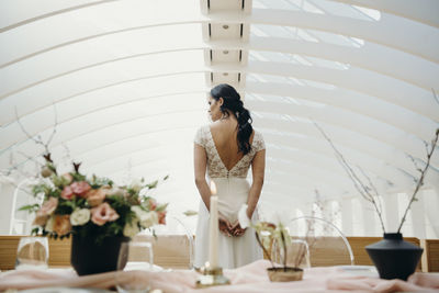 Back view of graceful young ethnic woman in white bridal dress standing alone near table setting for wedding ceremony in light hall