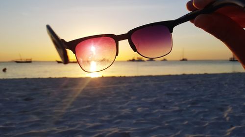 Close-up of person holding sunglasses on beach against sky during sunset