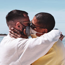 Gay couple wearing masks kissing against sky