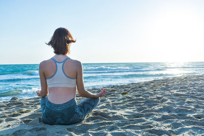 Rear view of woman meditating at beach against sky