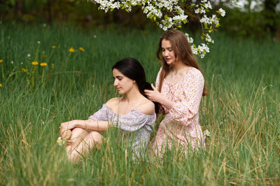 Two young girls in dresses are sitting under a white tree and braiding each other's hair