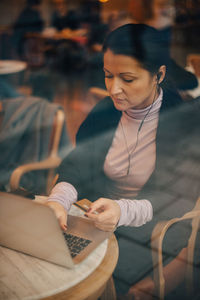 High angle view of businesswoman doing online shopping over laptop while sitting in cafe seen through window