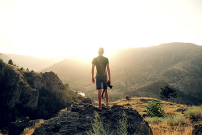 Rear view of man with camera looking at mountains while standing on rock against clear sky during sunset