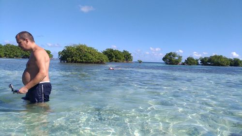 Side view of shirtless man standing in sea at beach against blue sky