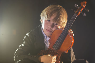 Boy holding violin while sitting against black background