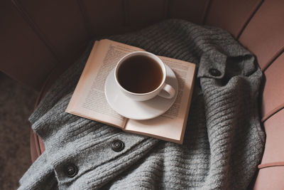 Mug of coffee with open paper book, knitted textile on cozy armchair at home closeup. good morning.