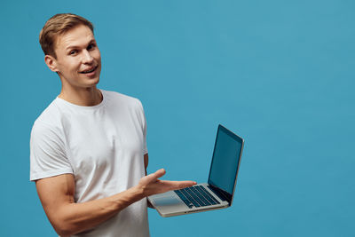 Portrait of young man using laptop while standing against blue background