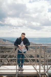 Young man standing on railing against sky