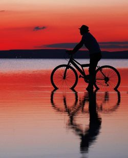 Silhouette man riding bicycle at sea against sky during sunset