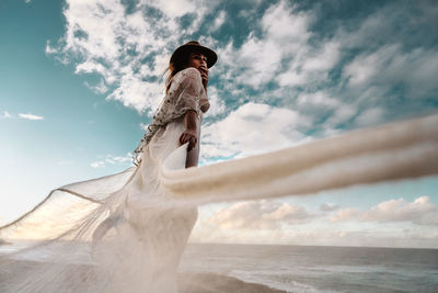 Low angle view of woman wearing white dress while standing at beach against cloudy sky during sunset