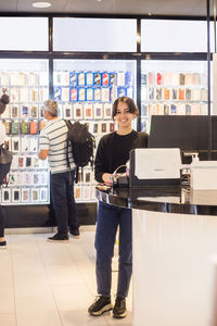 Full length portrait of confident female sales clerk standing by checkout against customers in shop