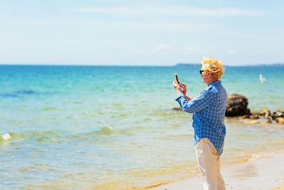 Elderly woman standing on the beach and taking photo on a mobile phone