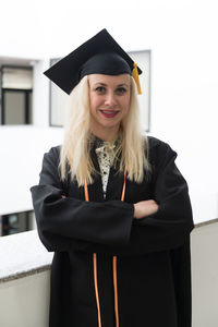 Young woman wearing graduation gown standing against wall