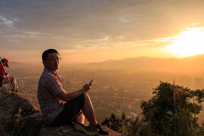 Portrait of man with smart phone sitting on mountain against cloudy sky during sunset