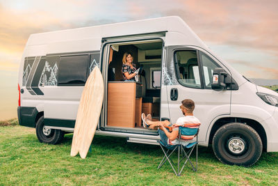 Woman drying pa in camper van while man playing ukulele outdoors