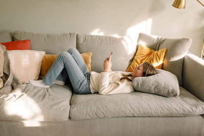 Full length side view of teenage girl lying on sofa surfing internet through smart phone at home