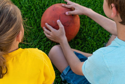 Unrecognizable children sitting on green grass and holding ball outdoors