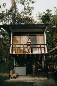 From below full body of young long haired female traveler in stylish swimwear standing on balcony of beach house located near green trees in summer day in uvita town in costa rica