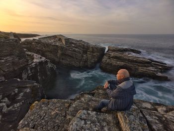 Man sitting on rock by sea against sky during sunset