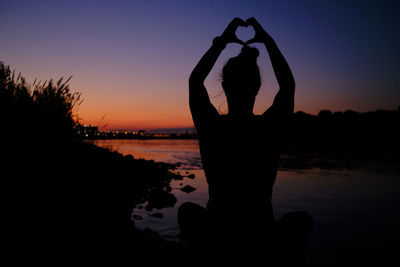 Young woman sitting at a river bench at sunset forming a heart over her head with her hands