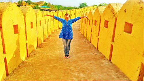 Full length of young woman with arms outstretched standing on footpath amidst walls