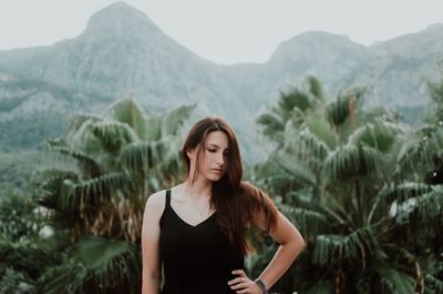 Worried young woman with hand on hip standing against trees and mountains