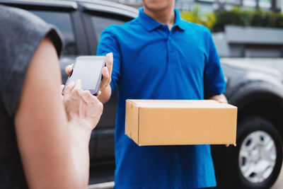 Midsection of woman holding mobile phone while delivery man holding box outdoors