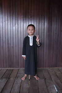 Portrait of boy showing peace sign while standing against wooden wall