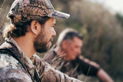 Side view of male hunter in camouflage jacket and cap sitting in grass and resting during hunting in autumn