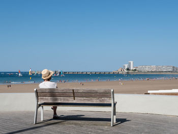 Rear view of woman sitting on bench by sea against clear sky