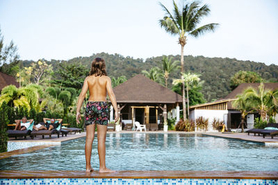 Rear view of shirtless girl standing at poolside in tourist resort during vacation