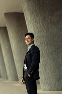 Side view of content handsome businessman standing with hand in pocket near building in city and looking away