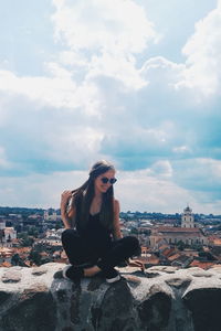 Young woman sitting on retaining wall against cityscape