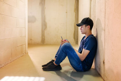 Side view of young man using phone while sitting on wall