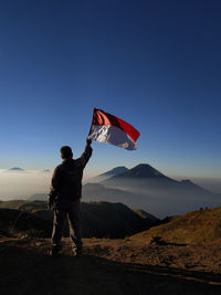 Rear view of man holding flag while standing on mountain against sky