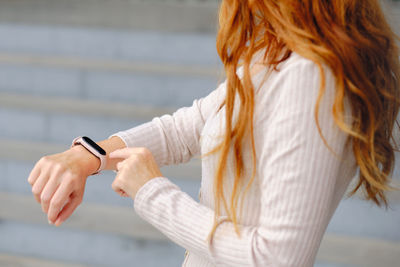Side view of crop red haired unrecognizable female in casual outfit standing on street and checking information on smart bracelet