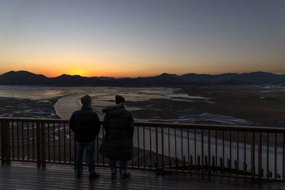 Rear view of people on shore against sky during sunset