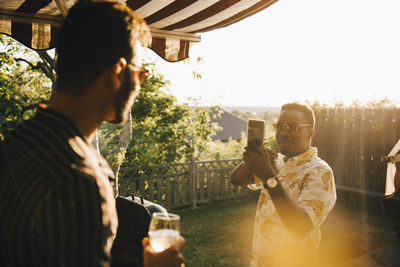 Man photographing friend with wineglass through smart phone at yard