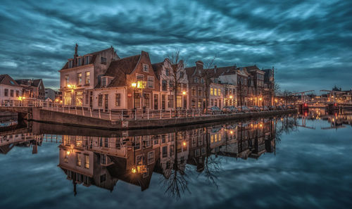 Illuminated buildings by river against cloudy sky at dusk