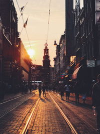 People walking on railroad tracks in city against sky during sunset
