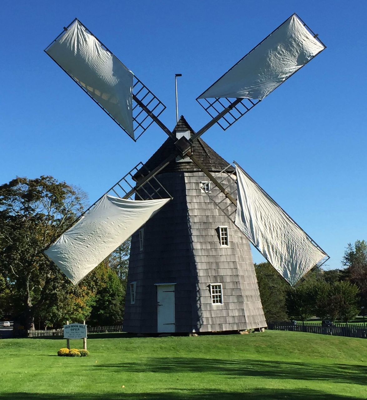 building exterior, architecture, built structure, grass, low angle view, wind power, alternative energy, clear sky, renewable energy, windmill, environmental conservation, sky, blue, traditional windmill, tree, tower, religion, sunlight, field, patriotism