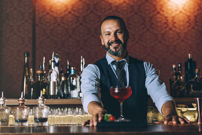 Portrait of smiling mature bartender standing by cocktail on bar counter