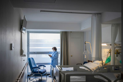 Elderly patient woman in robe and protective medical mask standing at window of hospital robe and browsing cellphone