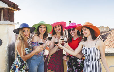 Portrait of smiling female friends toasting drinks while standing in balcony