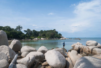 Rear view of man standing rocks by sea against blue sky